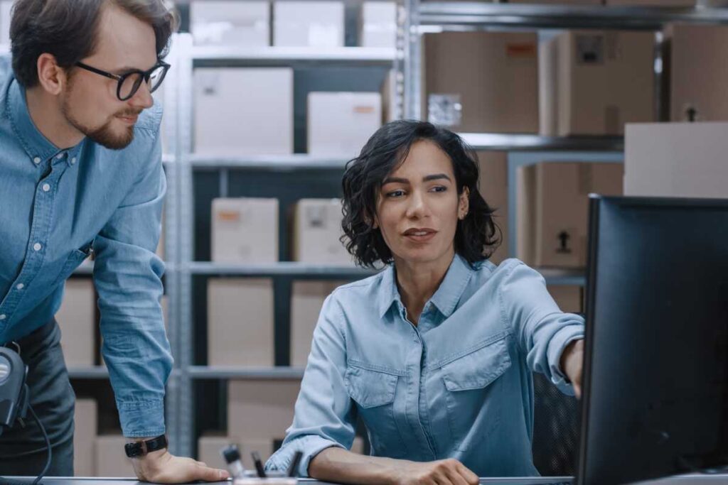 Male and Female Warehouse Inventory Managers Talking, Using Personal Computer and Checking Stock. Female Pointing at Screen. In the Background Rows of Shelves Full of Cardboard Box Packages.