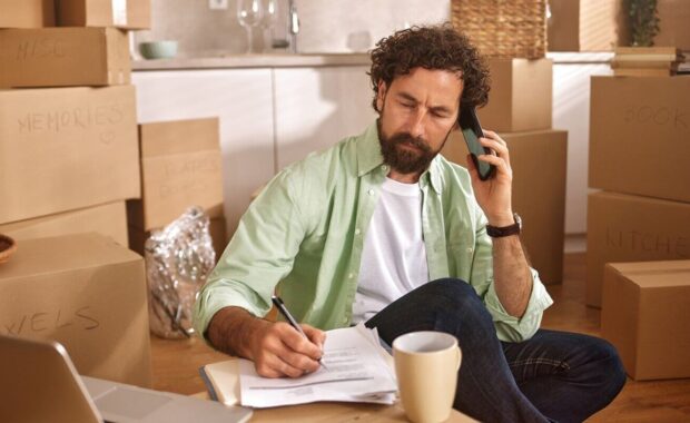 man sits on the floor of his new home, taking a phone call while writing notes