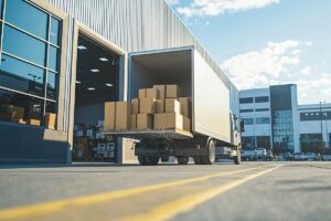 Delivery truck unloading boxes at warehouse dock under bright blue sky.