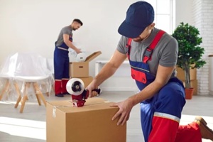 Mover sealing a cardboard box with packing tape while another mover prepares items in the background.